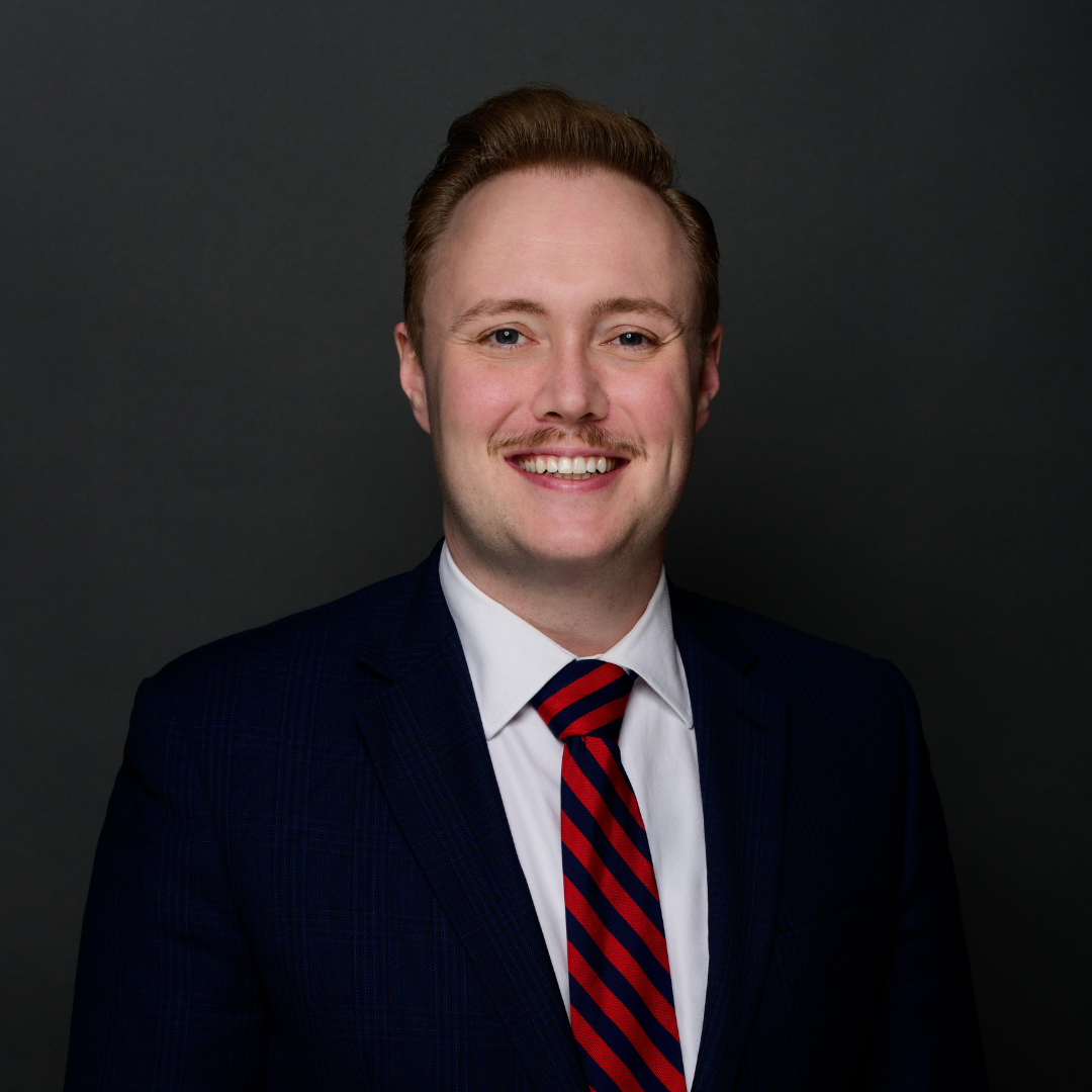 Smiling man in a navy suit and red-and-blue striped tie, head-and-shoulders portrait against a dark background.